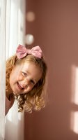 Adorable young girl with curly blonde hair and pink bow peeking around white door frame with joyful smile in warm sunlit interior.