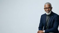 Senior African American male professional with gray beard and glasses wearing navy blazer and gray shirt, isolated on white background for corporate portraits, business headshots, and executive leadership content.
