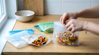 Young adult Caucasian female hand sealing a clear plastic food storage container filled with colorful mixed vegetables on wooden kitchen counter with additional storage containers and kitchenware visible, demonstrating food preservation and meal prep organization.