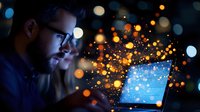 Young bearded Caucasian man in glasses working on laptop at night with magical glowing particles and bokeh lights creating ethereal atmosphere. Creative digital concept.