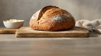 Freshly baked artisan sourdough bread loaf on wooden cutting board with flour bowl, rustic kitchen setting for bakery and homemade cooking concepts.