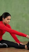 Young Hispanic woman in red athletic jacket stretching on wooden floor during fitness workout routine against textured green wall background.