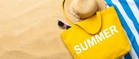 Yellow summer tote bag with straw hat, sunglasses, and striped towel on sandy beach, flat lay top view for seasonal vacation concepts.