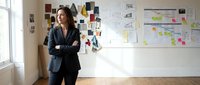 Woman in business casual attire standing confidently with crossed arms in front of mood boards and design sketches on wall, suitable for creative industry, startup, and professional workspace concepts.