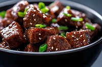 Glazed beef chunks with sesame seeds and green onions in dark bowl, close up view of Asian style meat dish with sweet soy sauce marinade.