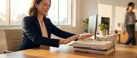Professional businesswoman using printer in modern office workspace with natural lighting and colleague in background.