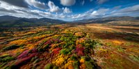 Dramatic autumn landscape with vibrant red, orange, and yellow foliage stretching across rolling hills under dynamic cloudy blue sky.