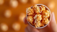 Caramel popcorn in paper cup held by person against blurred orange background with bokeh lights, sweet snack for movie night or party.