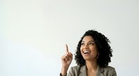 Woman with curly dark hair and olive skin tone wearing beige blazer, pointing upward with index finger and smiling, looking up. Suitable for business, ideas, and motivation concepts.