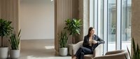 Young professional woman working on laptop in modern office lobby with natural light and plants creating productive workspace atmosphere.