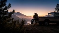 Man sitting on vintage pickup truck bed at sunrise overlooking misty mountain valley and river. Outdoor adventure, travel, and camping lifestyle scene for editorial and lifestyle use.