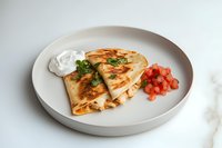 Mexican quesadilla with melted cheese, diced tomatoes and sour cream served on round white plate, garnished with fresh cilantro against light background.