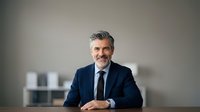 Middle-aged man with salt-and-pepper hair and facial hair, Caucasian ethnicity, wearing dark business suit and tie, seated at desk in modern office environment with blurred background, professional corporate portrait.