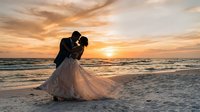 Romantic couple kissing on beach at sunset during wedding ceremony with golden sky and ocean waves creating dreamy atmosphere for love celebration.