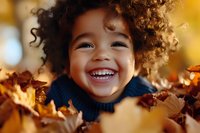Joyful African American child with curly hair laughing while playing in autumn leaves, wearing navy sweater. Natural golden bokeh background creates warm seasonal mood.