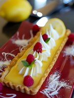 Lemon tart with fresh raspberries and whipped cream swirls on red plate, garnished with mint leaves and dusted with powdered sugar, close up view of French pastry.