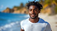 Young man with dark curly hair and medium brown skin tone wearing white t-shirt smiling at camera on sunny beach with blue ocean and sandy shore in background.