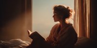 Young woman in bathrobe reading book by window at sunrise, warm golden light creating peaceful morning atmosphere for relaxation and self-care.
