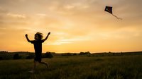Child flying kite in open field at golden hour sunset, arms raised in joy and freedom, outdoor recreational activity for leisure and happiness.