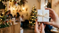 Person holding wrapped gift box with gold ribbon at festive holiday gathering with decorated trees and warm lights.