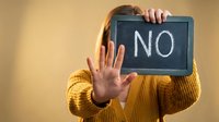 Young woman in yellow sweater holding chalkboard with NO text while making stop gesture with hand for rejection and refusal concepts.