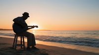 Silhouetted musician playing acoustic guitar on sandy beach at sunset, seated on stool facing ocean waves, golden hour coastal scene for music and lifestyle content.