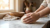 Baker kneading fresh dough on floured counter in warm kitchen with natural sunlight streaming through window creating cozy homemade bread making atmosphere.