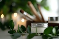 Natural cosmetic cream jar surrounded by fresh green leaves and cinnamon sticks on white surface with soft bokeh lights, creating spa-like atmosphere for beauty product photography.