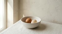 Minimalist white ceramic bowl with fresh brown eggs on linen tablecloth near window with natural sunlight creating soft shadows on textured wall.