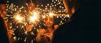 Hands holding lit sparklers with glowing sparks bursting against dark background, celebrating festive occasions and New Year's Eve parties.
