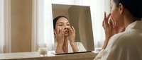 Young woman with East Asian ethnicity examining her face in mirror, touching cheeks and skin while wearing white bathrobe in bright bathroom. Skincare routine and beauty concept for cosmetics and wellness.