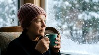 Senior woman in knit hat drinking hot beverage by snowy window during winter storm, cozy indoor warmth contrasting cold weather outside.