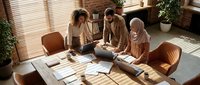 Three diverse business professionals collaborating around modern conference table in bright office with brick walls and natural lighting.
