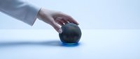 Hand touching a dark round smart speaker with glowing blue light on a clean light blue background, modern home technology concept.