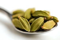 Aromatic green cardamom pods in silver spoon on white background, macro shot showing detailed texture of spice used in Indian and Middle Eastern cuisine.
