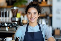 Cheerful young Hispanic female barista wearing apron smiles warmly at cafe counter. Natural lighting highlights genuine expression and professional service atmosphere.