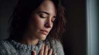 Young woman with closed eyes in gray sweater by window, contemplative expression in moody lighting, conveying solitude and introspection.