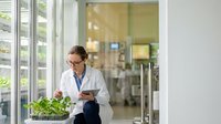 Female scientist in white lab coat examining green plants in modern research facility with tablet computer for agricultural biotechnology studies.