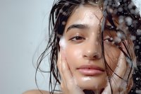 Young Hispanic woman with wet hair and glowing skin behind water drops on glass, beauty portrait showcasing natural skincare and hydration concept for cosmetic advertising.
