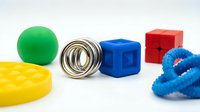 Colorful sensory toys including green ball, yellow textured roller, metal spring coil, blue cube, red block, and blue spiky fidget toy on white background for child development and tactile play.