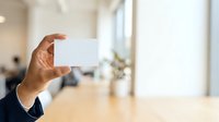 Close-up of male hand holding blank white business card in modern office environment with blurred background, suitable for corporate branding and networking concepts.