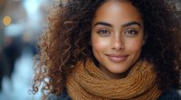 Young African American woman with natural curly hair smiling warmly while wearing cozy knit scarf on autumn day. Soft bokeh background creates welcoming atmosphere.