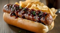 Grilled sausage in soft bun topped with caramelized onions and herbs, served with potato chips on rustic wooden surface. Dark moody food photography.