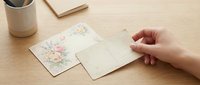 Woman's hand opening blank greeting card with floral design on wooden desk next to notebook and plant pot, stationery mockup for invitations and correspondence.