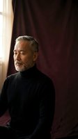 Mature Caucasian man with gray beard in black turtleneck sitting peacefully by window with burgundy curtains in elegant interior setting.