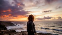 Young woman with long brown hair and fair complexion standing on rocky coastline at sunset, gazing toward ocean horizon. Dramatic golden and purple sky with clouds. Waves crashing on rocks below. Contemplative mood, solitude, travel, nature.