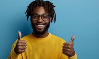 Young African American man with dreadlocks and glasses showing thumbs up, wearing yellow sweater against blue background. Genuine cheerful expression portrays positivity.