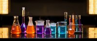 Colorful chemical liquids in laboratory glassware, flasks and beakers with vibrant orange, purple and blue solutions on reflective surface.