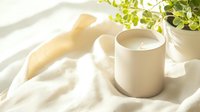 White ceramic cup with fresh milk on soft beige linen fabric in morning sunlight with green plant leaves, creating minimalist composition for lifestyle and wellness.
