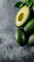 Fresh ripe avocados with one cut in half showing creamy flesh and pit cavity on gray concrete background with green leaves, natural light food photography.